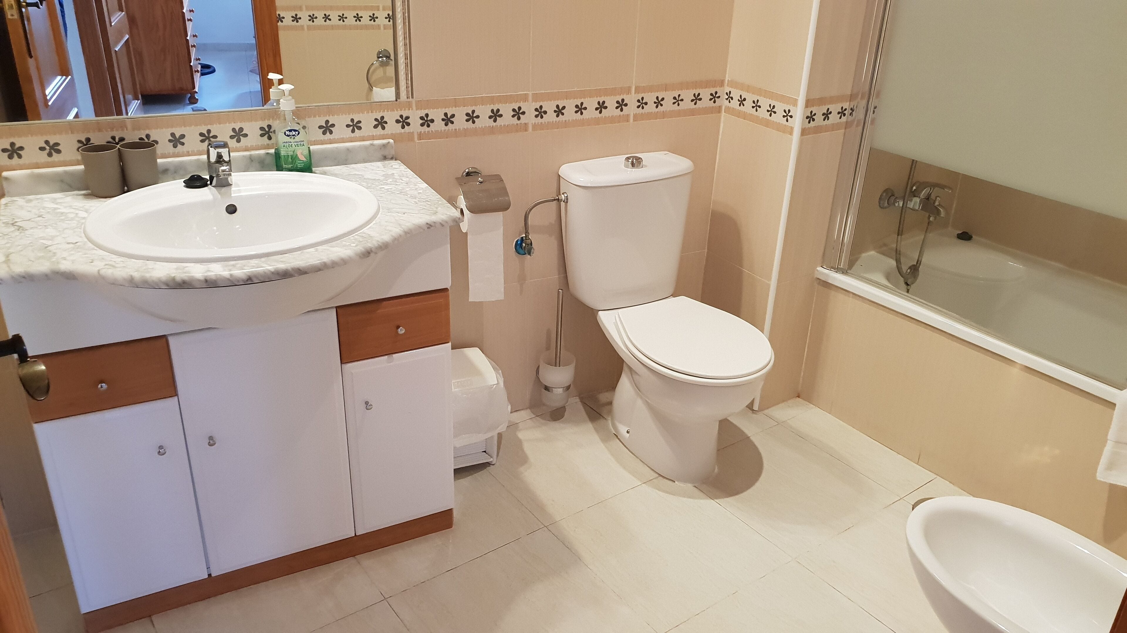 Un baño con lavamanos, inodoro y bidé blancos. Hay un espejo sobre el lavamanos, y las paredes están revestidas con un estampado floral.