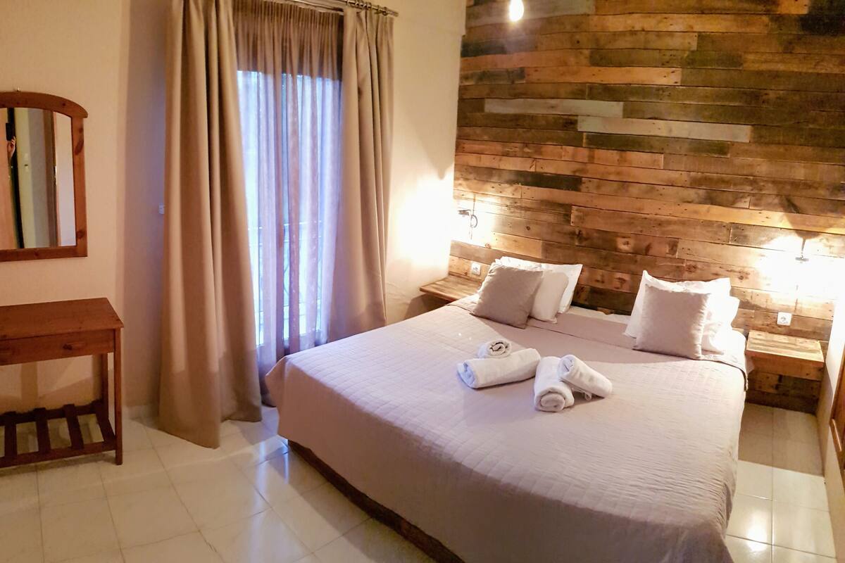 A hotel room with a large bed, wooden headboard, two bedside tables, a mirror, and a window with curtains.