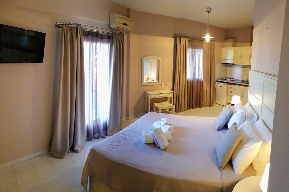 A hotel room with a large bed, a bedside table, a mirror, a small desk, and a kitchenette.