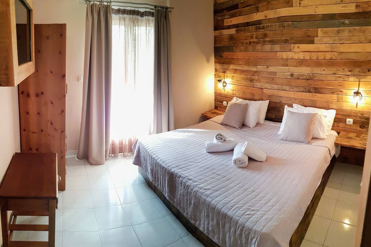 A bedroom with a wooden headboard, a bed with white bedding, a bedside table with a lamp, and a window with curtains.
