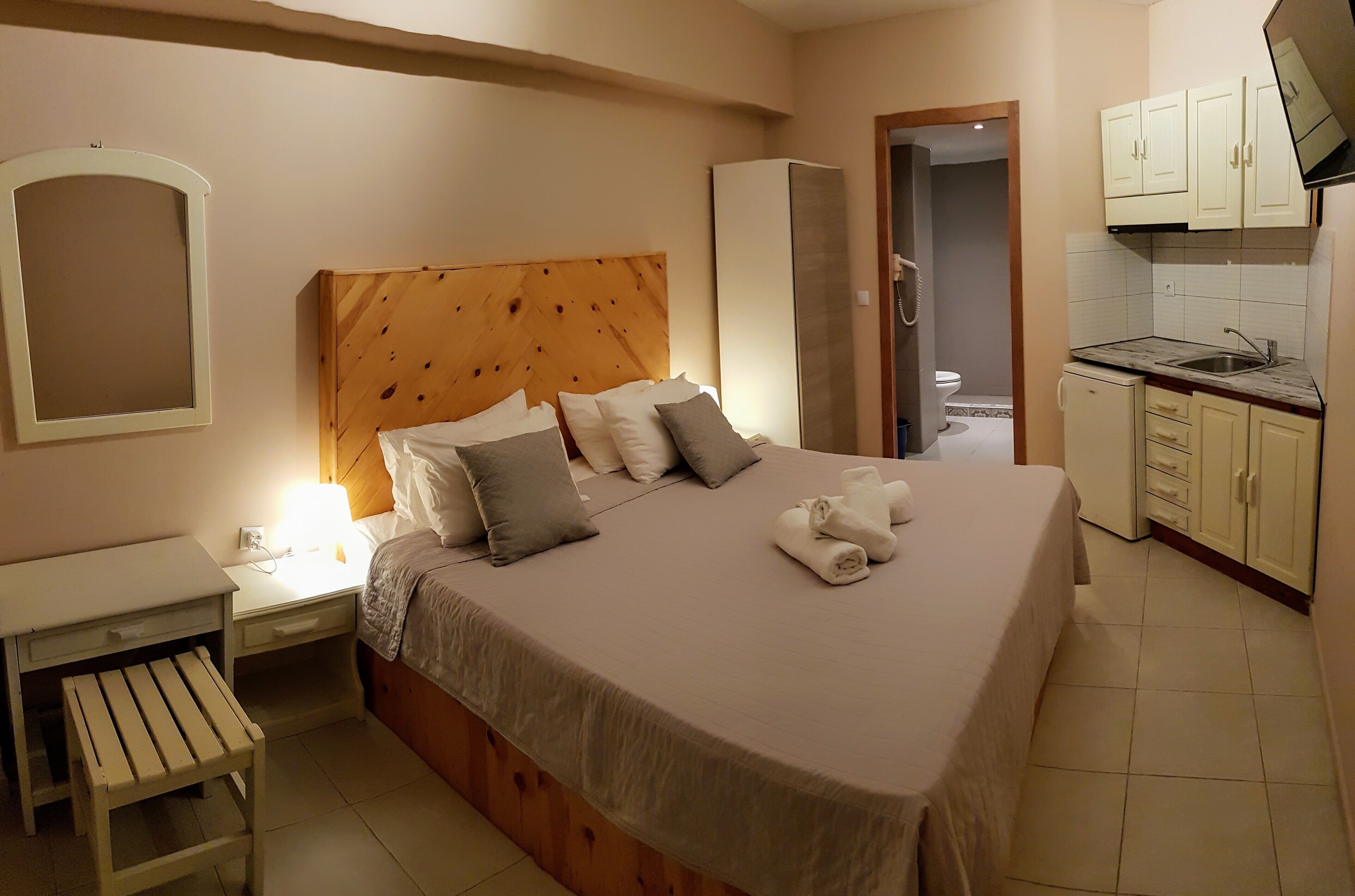 A hotel room with a bed, bedside table, lamp, and a small stool. There is a kitchenette with a sink and cabinets.