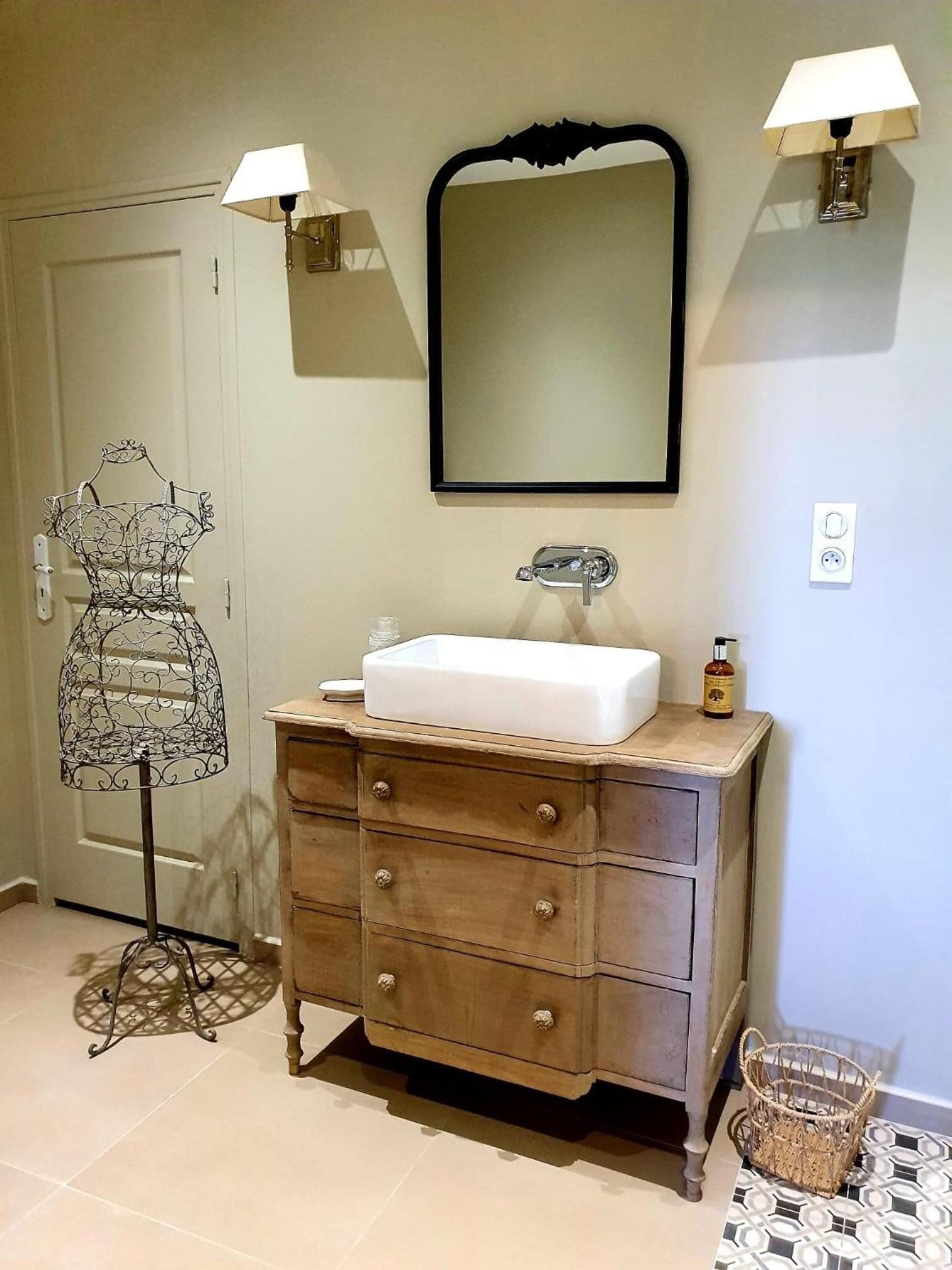 Baño con tocador de madera, un lavamanos blanco, un espejo, dos lámparas de pared, una botella de jabón para manos y un estante decorativo de metal.