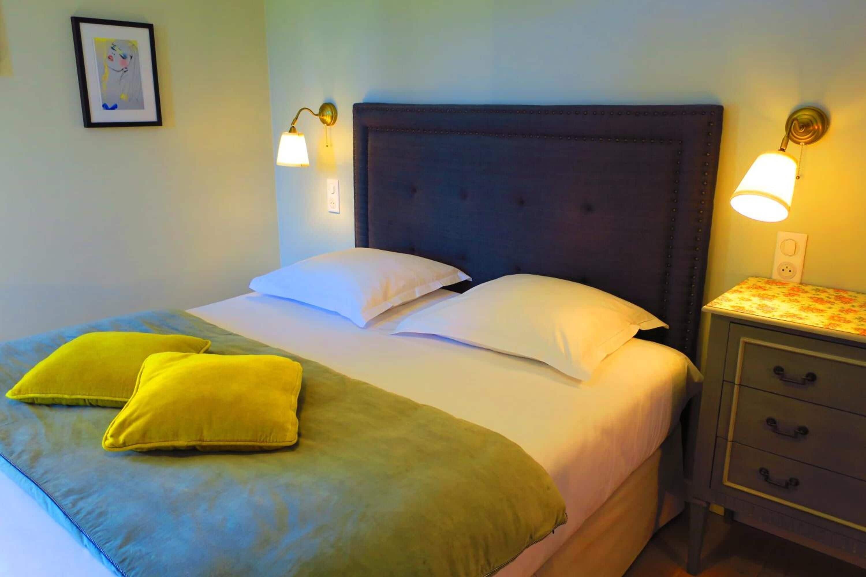 A neatly made bed with a dark headboard, two pillows, and a green throw blanket. There are two wall-mounted lamps on either side of the bed.