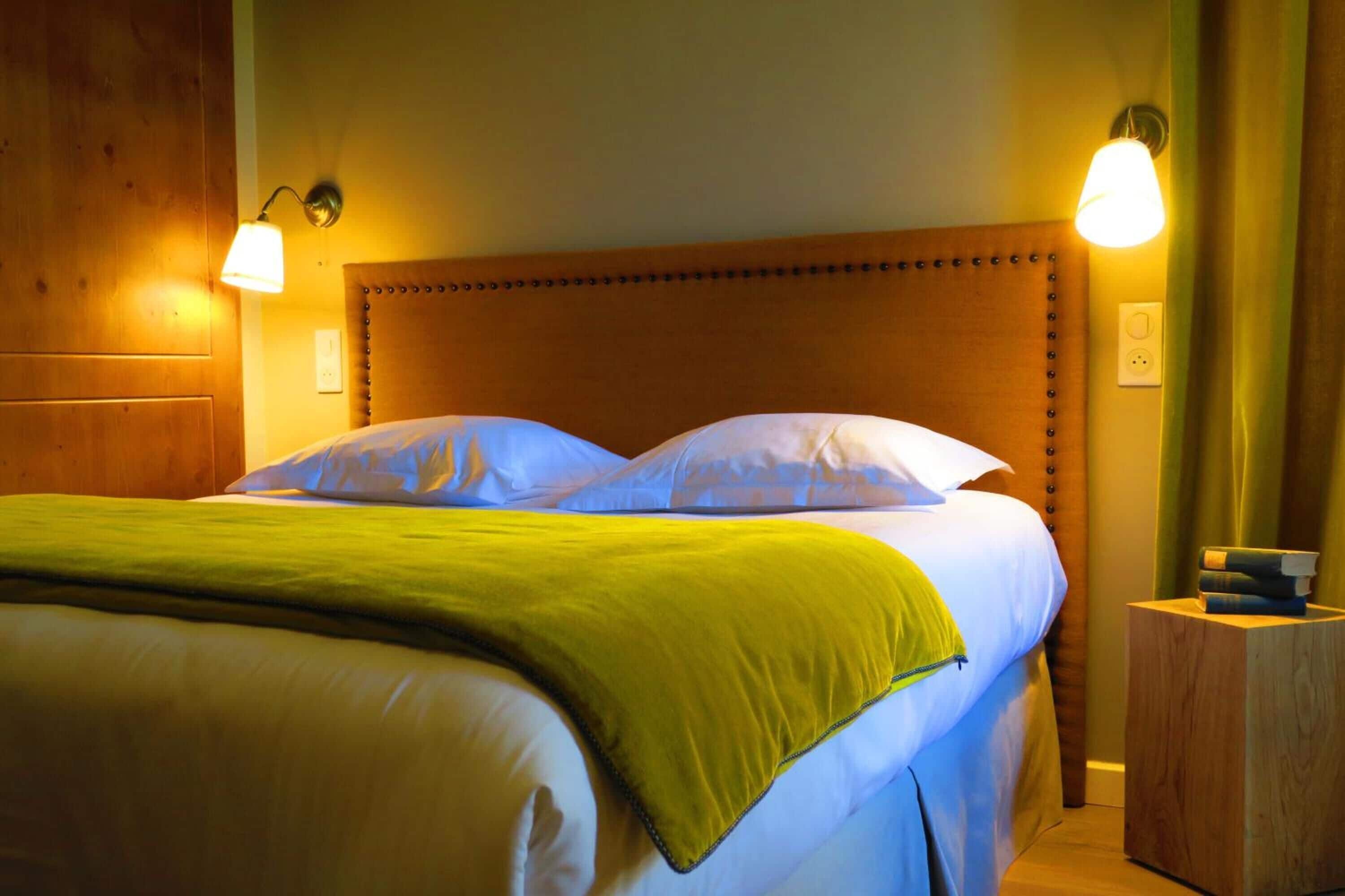 A neatly made bed with a green blanket and two pillows, a wooden headboard, and two wall-mounted reading lamps.