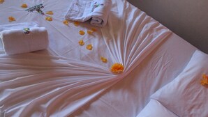 Una cama bien hecha con sábanas blancas, una toalla doblada y pétalos de flores amarillas esparcidos.