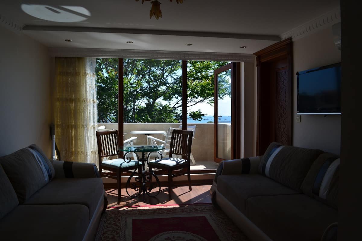 A living room with two sofas, a coffee table, and chairs. There is a large window with a view of trees and a balcony.