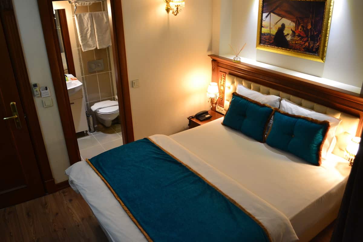 A hotel room with a bed, a nightstand, a lamp, a painting on the wall, and a bathroom visible through an open door.