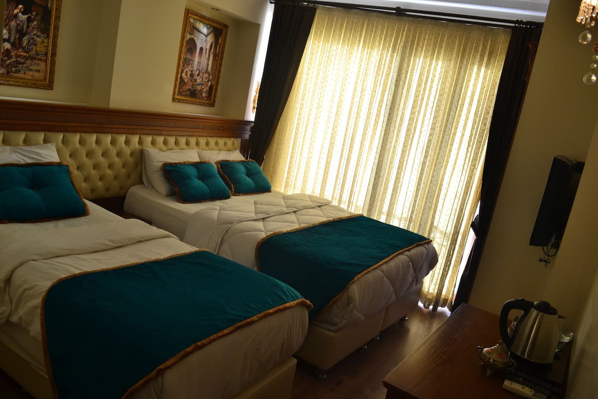 A hotel room with two beds, each with teal bedding and a wooden headboard. There is a wooden nightstand with a kettle and a small decorative item. The room has a large window with curtains, and artwork is visible on the walls.