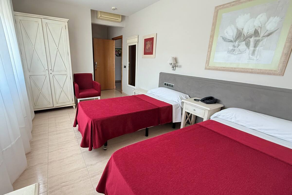 Quarto de hotel com duas camas de solteiro, uma cadeira vermelha, uma escrivaninha com telefone e um guarda-roupa.