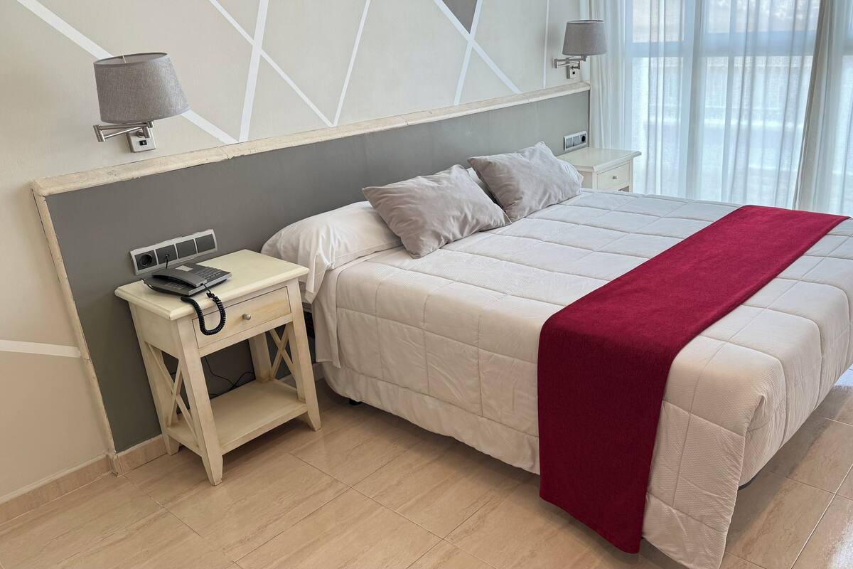 Quarto de hotel com cama, mesa de cabeceira, luminária e vista para o exterior pela janela.