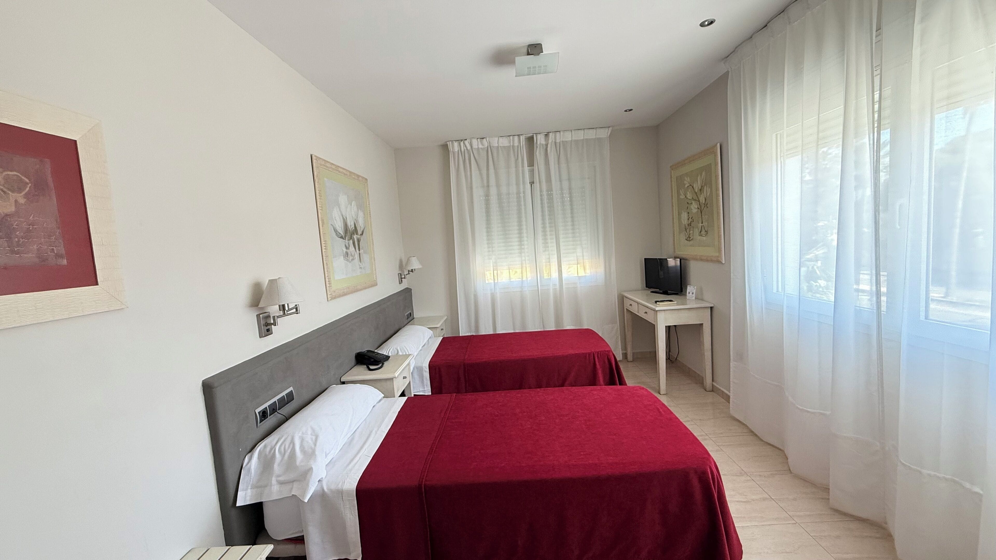 A hotel room with two beds, a desk with a TV, and a window with curtains.