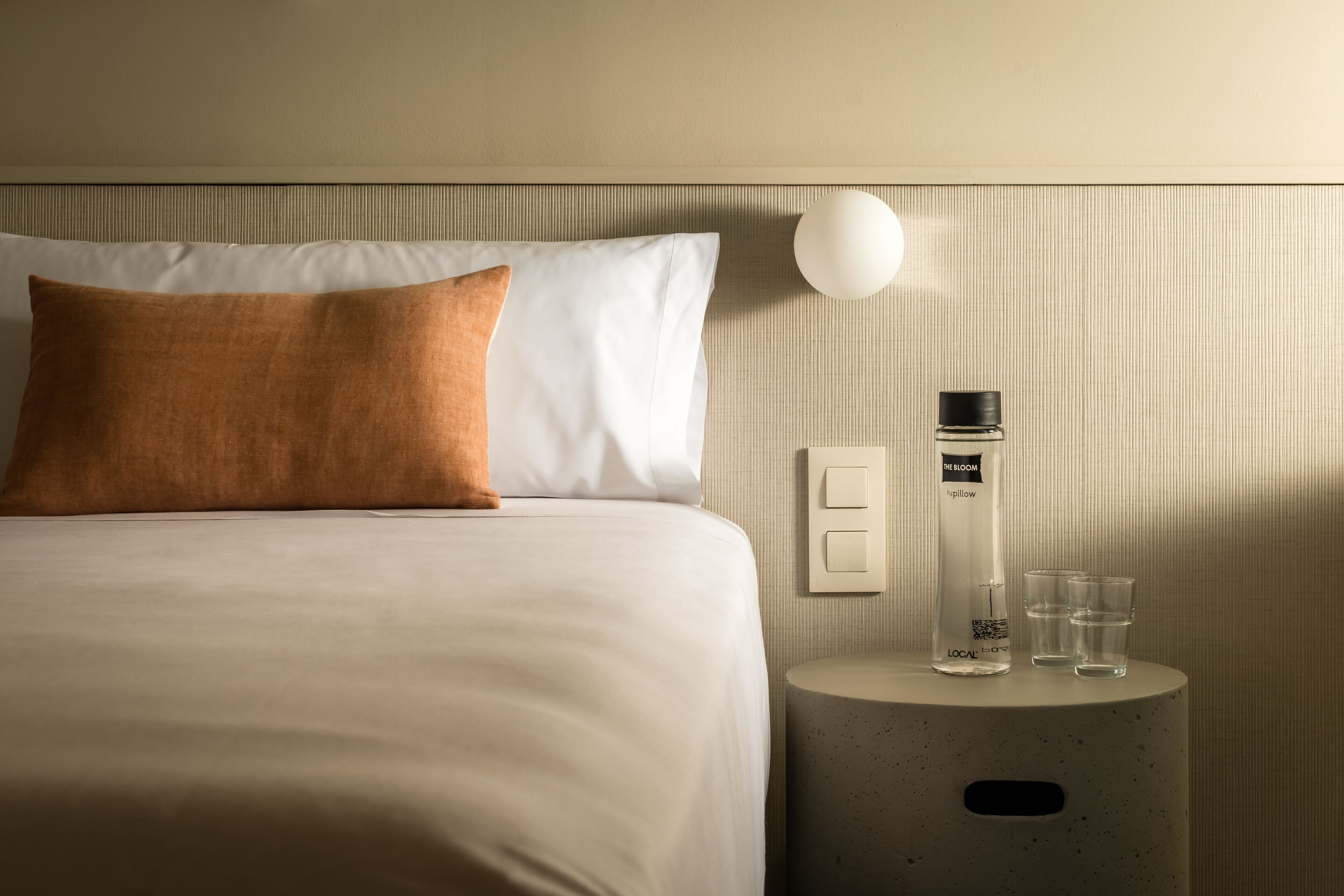 A neatly made bed with white and brown pillows, a bedside table with a bottle and glasses, and a wall-mounted light.