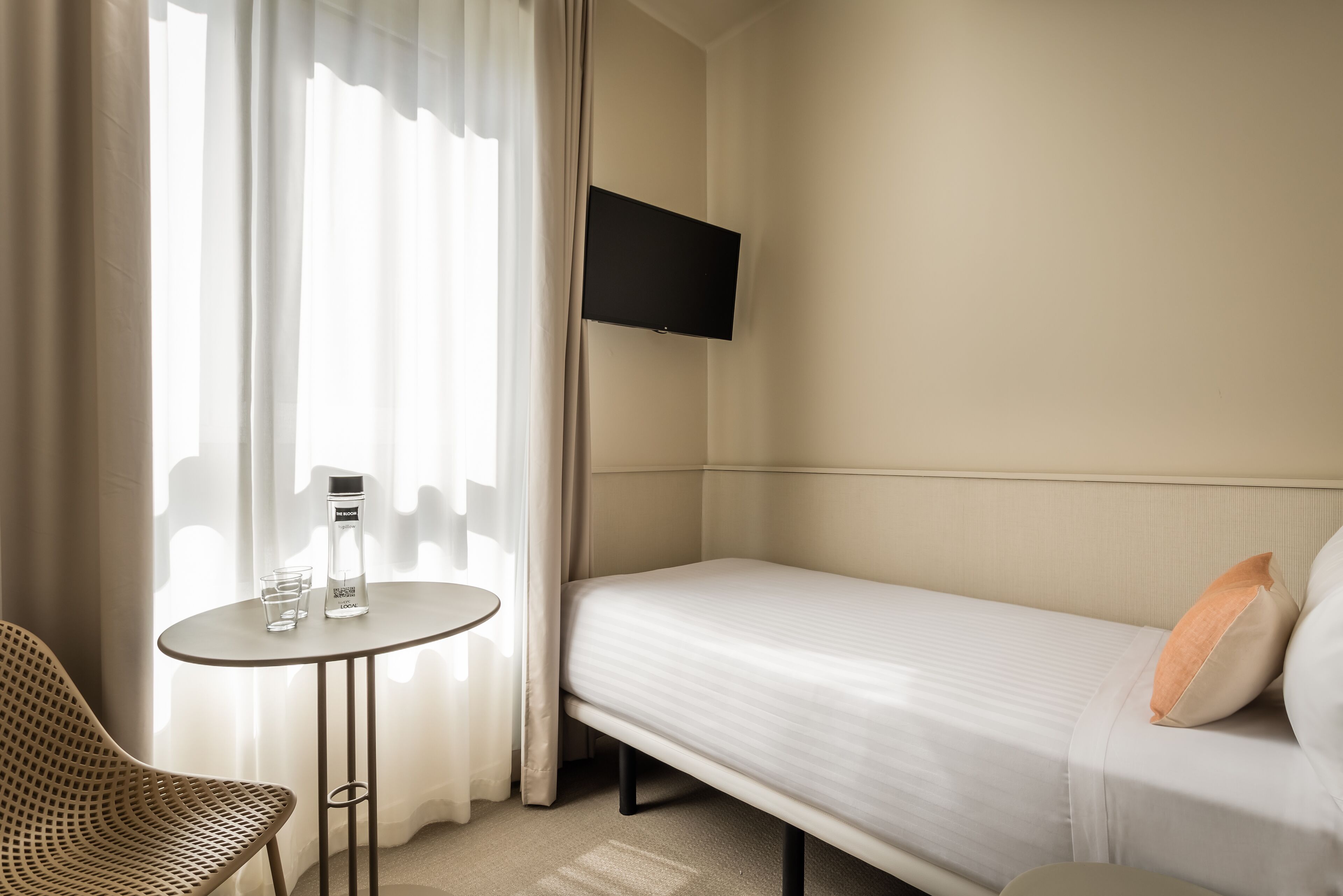 A hotel room with a bed, a TV mounted on the wall, a small table with a bottle and glass, and a chair.