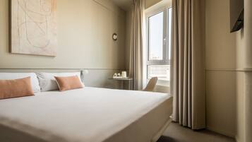 A hotel room with a large bed, a small table with a lamp, a chair, and a window with curtains.