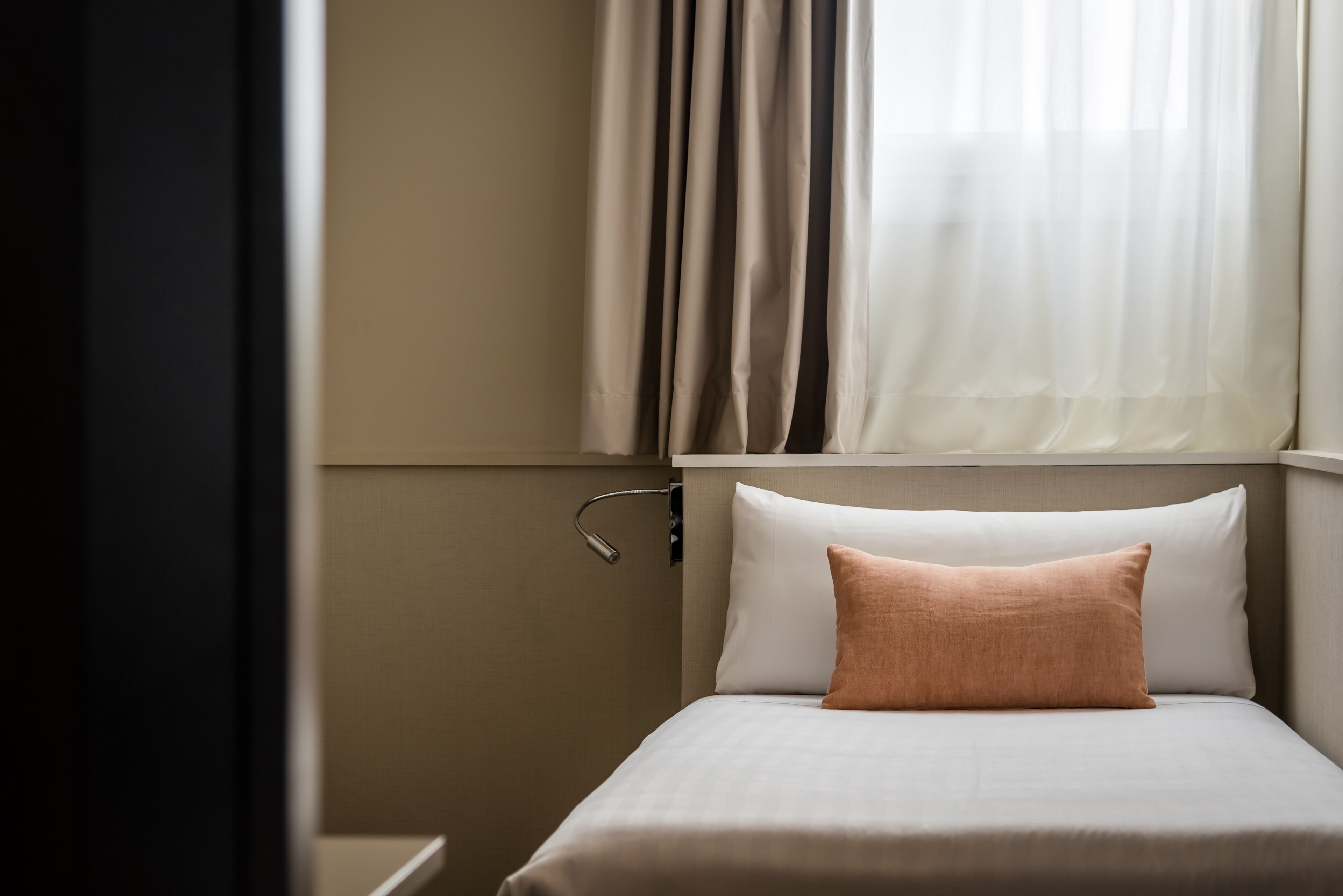 A neatly made bed with a single pillow, a bedside table, and a window with sheer curtains.