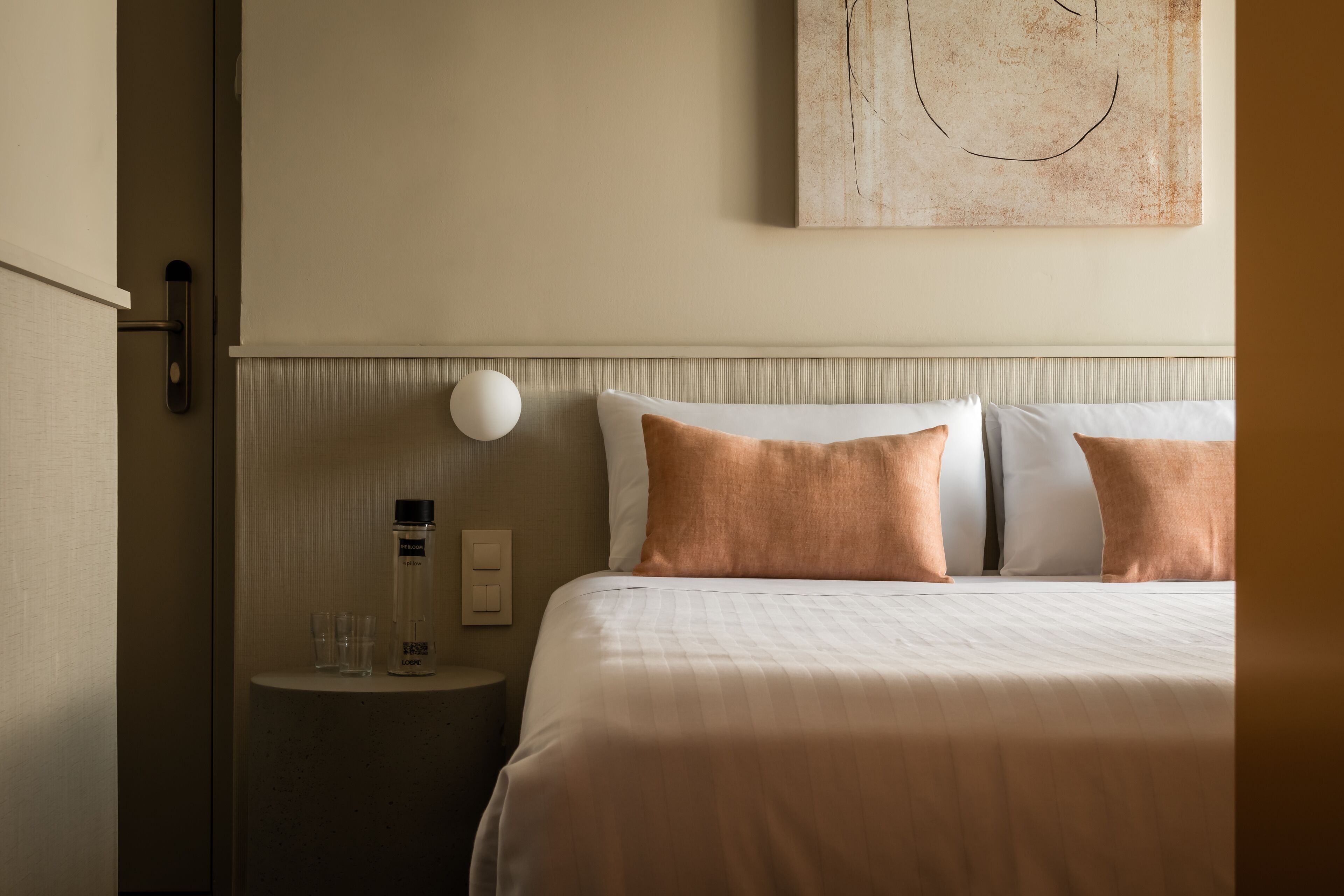Een net opgemaakt bed met twee kussens, een nachtkastje met een lamp, en een kunstwerk aan de muur.
