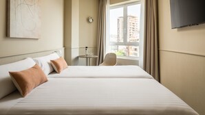 Habitación de hotel con dos camas, televisión, ventana con cortinas y vistas a edificios.