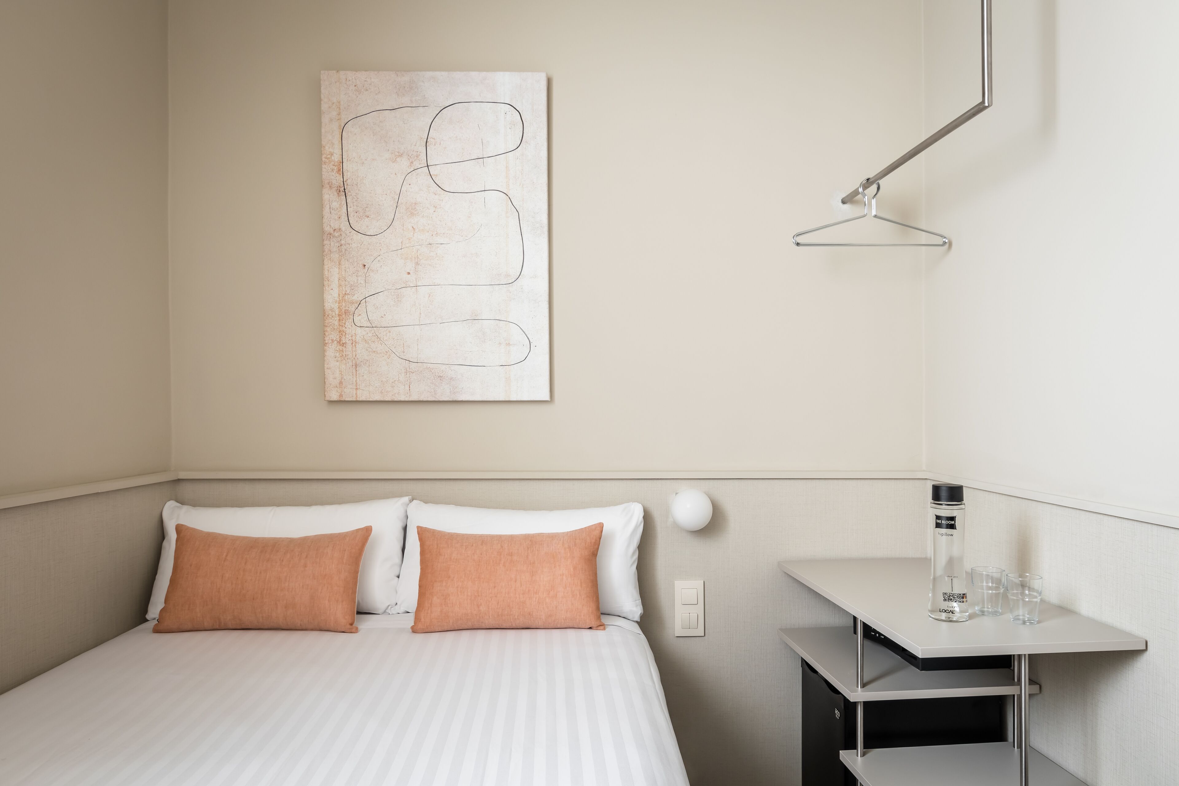 Une chambre d’hôtel bien aménagée, avec un lit, deux oreillers, une œuvre d’art abstraite accrochée au mur, une table de chevet avec une bouteille et des verres, et un luminaire suspendu.