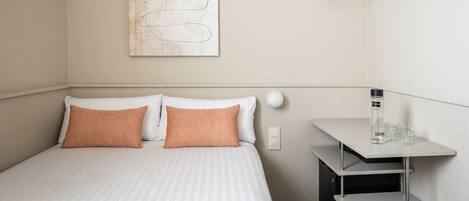 A neatly arranged hotel room with a bed, two pillows, a wall-mounted abstract artwork, a bedside table with a bottle and glasses, and a hanging light fixture.