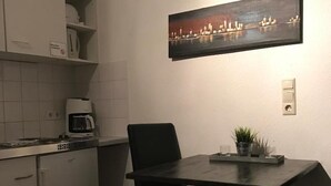 Una cocina compacta con microondas, cafetera y un refrigerador pequeño. Un comedor con dos sillas y una planta en maceta se encuentra debajo de una obra de arte en la pared que representa un paisaje urbano.