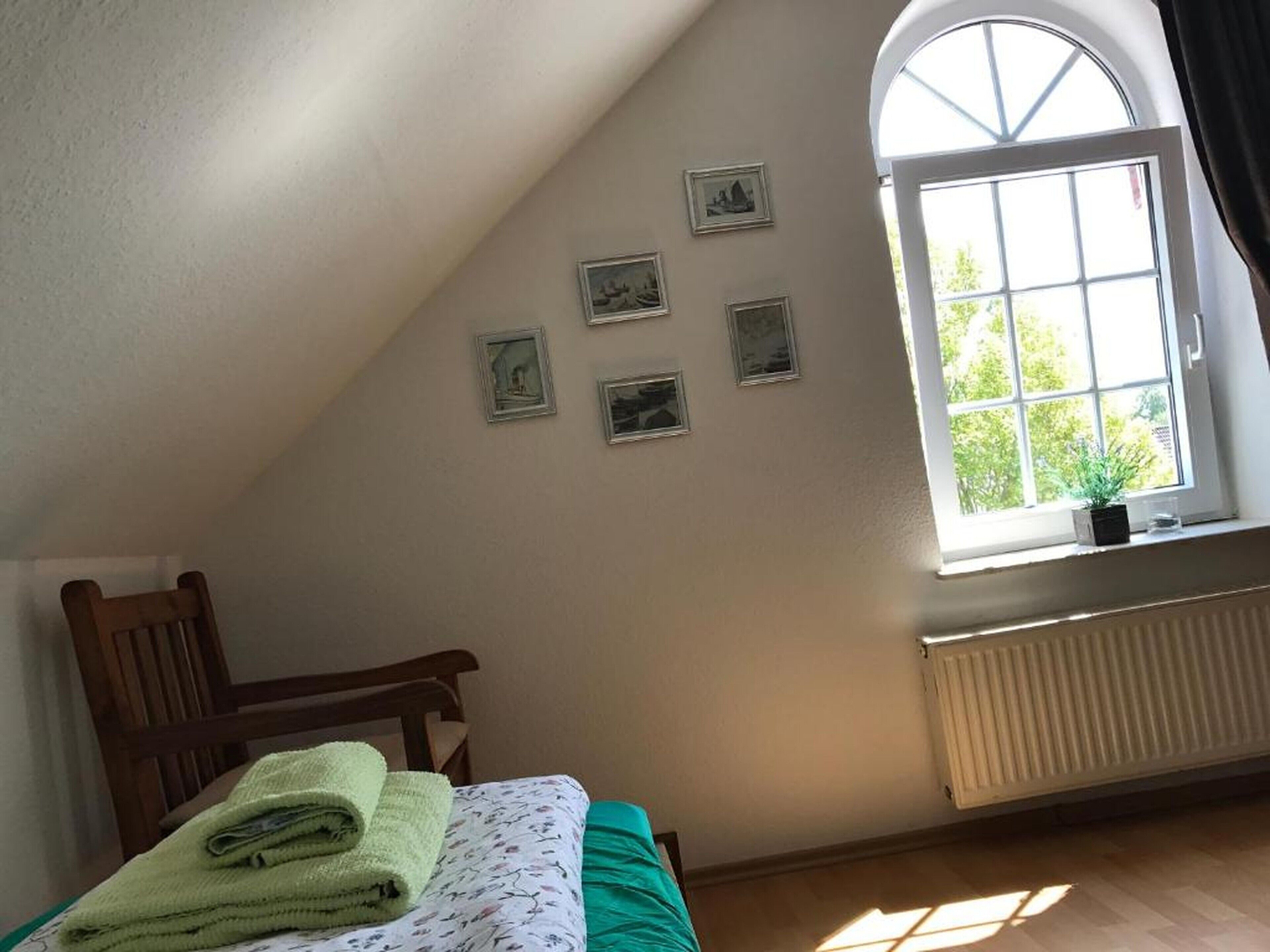 A bedroom with a sloped ceiling, a bed with a green blanket, a wooden chair, a window with a view of greenery, and a radiator.