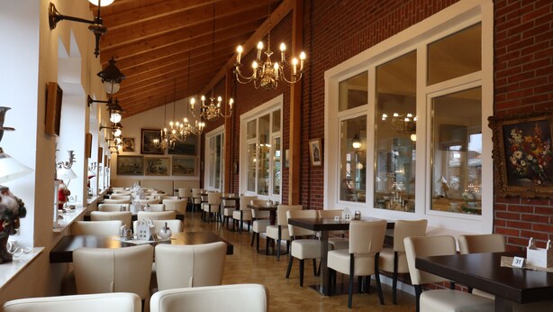 Un restaurante con varias mesas y sillas, ventanas amplias y candelabros colgantes.