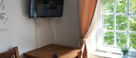 A room with a wooden table, a chair, a TV mounted on the wall, and a window with curtains.
