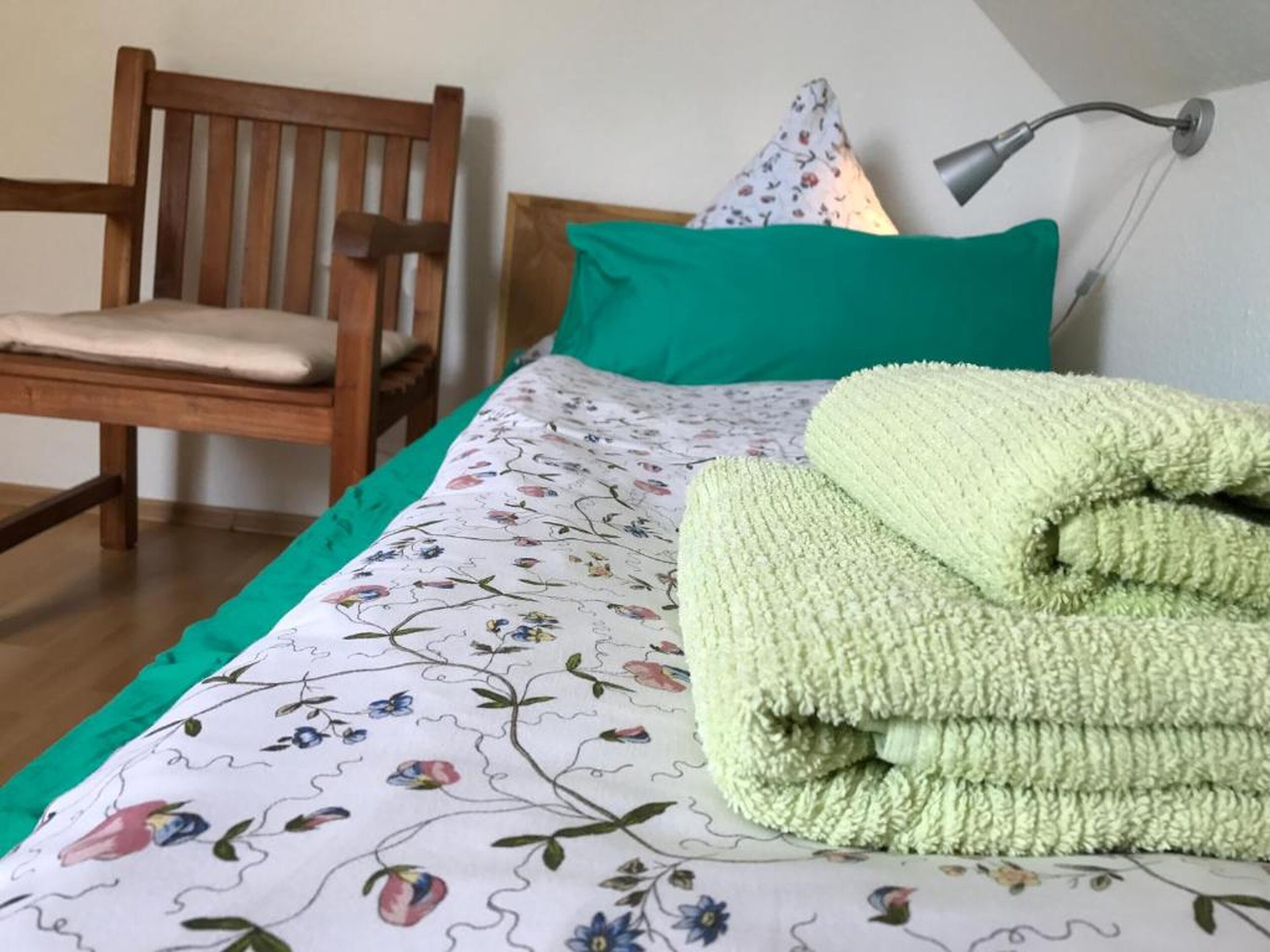 Ein Bett mit floralem Überwurf, einem grünen Kopfkissen und einem gefalteten grünen Handtuch.