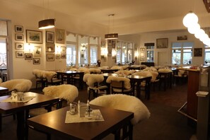 Ein Restaurant mit mehreren Tischen und Stühlen, gerahmten Bildern an den Wänden und großen Fenstern, die für natürliches Licht sorgen.