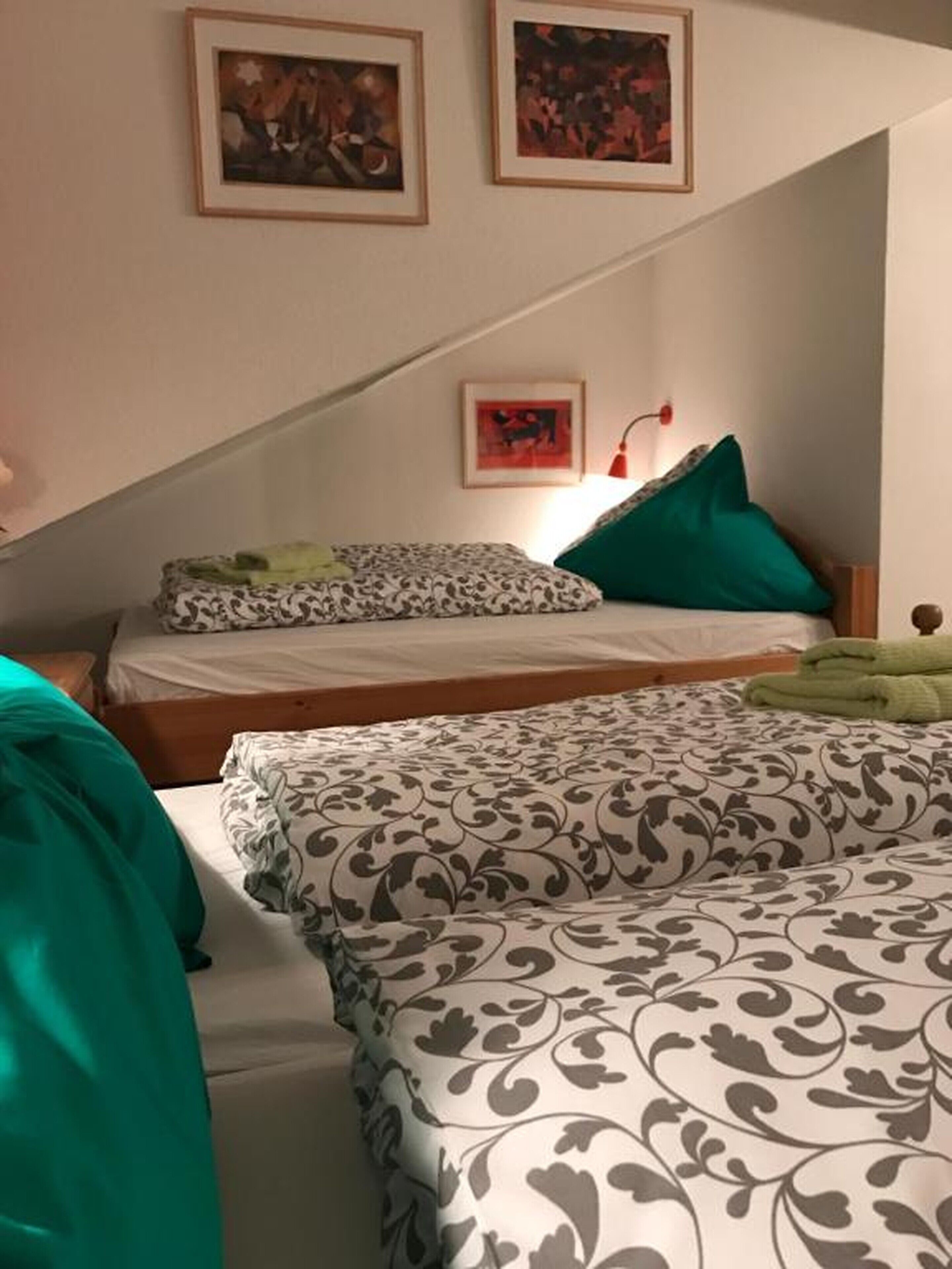 Un dormitorio con dos camas, una con cubrecama estampado y la otra con un diseño liso. Hay dos cuadros en la pared, una lámpara roja y un cojín verde en una de las camas.