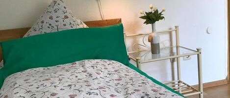 Een slaapkamer met een bed, een nachtkastje met een lamp, een vaas met bloemen, en een wand met een bloemenpatroon en hartversieringen.