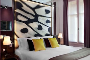 Quarto de hotel com uma cama grande, duas luminárias de cabeceira, uma TV na parede e uma janela com cortinas.