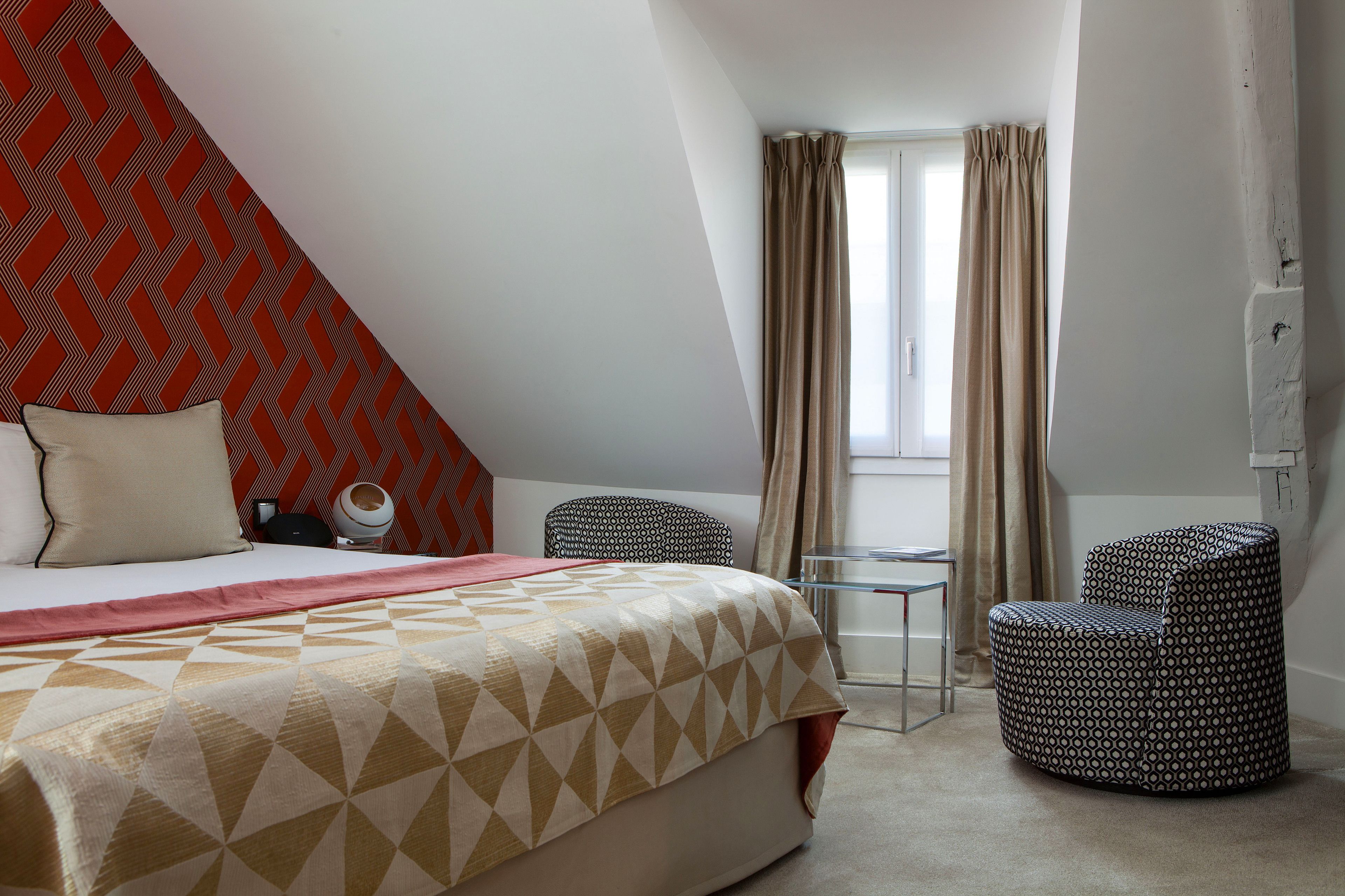 Une chambre d’hôtel avec un lit, deux chaises, une petite table et une fenêtre avec des rideaux.