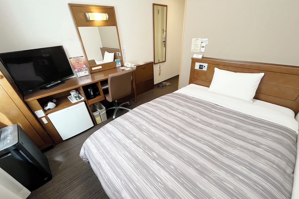 Une chambre d’hôtel avec un lit, un bureau avec une télévision, un miroir et un petit réfrigérateur.