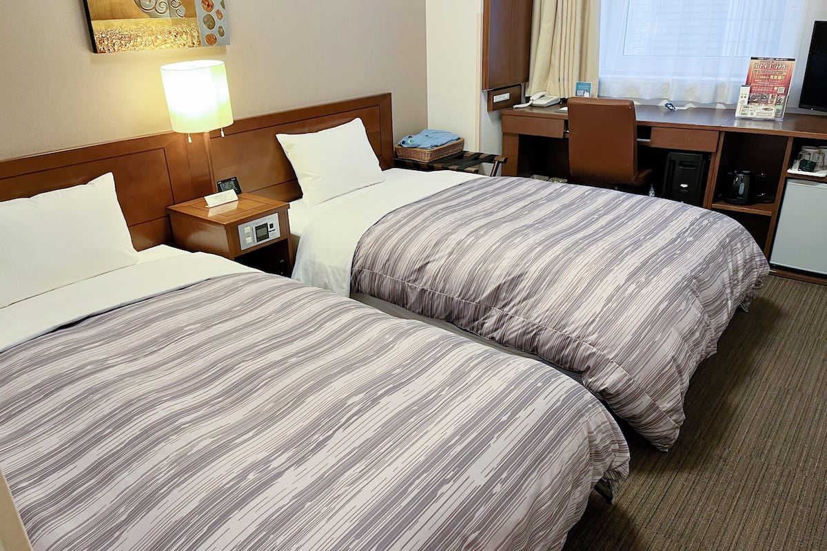 Une chambre d’hôtel avec deux lits, un bureau, une chaise, une lampe, un téléphone et une petite table avec un panier.