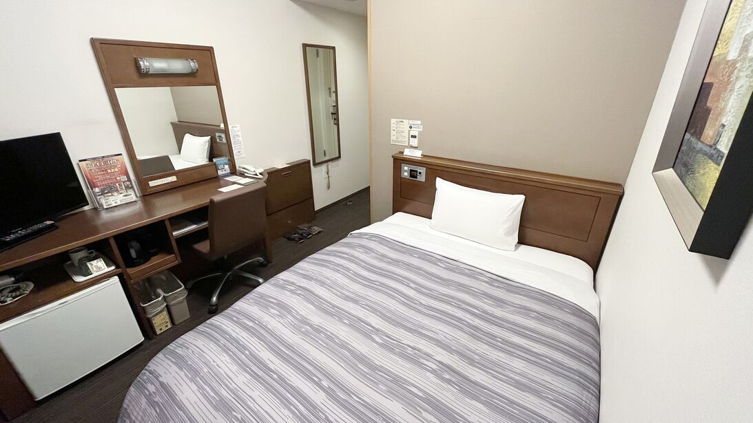 A hotel room with a bed, a desk with a computer, a mirror, a small fridge, and a bedside table.