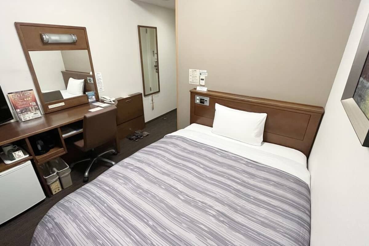 Une chambre d’hôtel comprenant un lit, un bureau avec un ordinateur, un miroir, un petit réfrigérateur et une table de chevet.