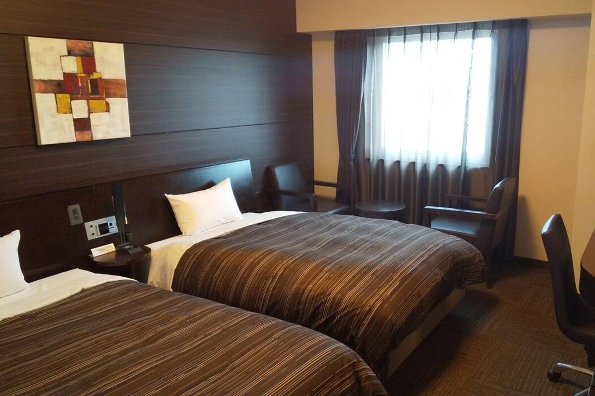 A hotel room with two beds, a desk with a television, a chair, and a window with curtains.