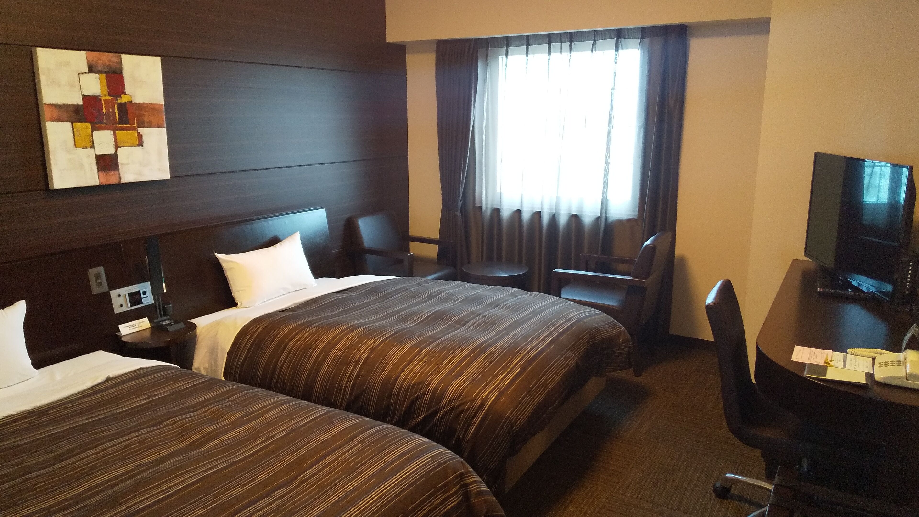 A hotel room with two beds, a desk with a television, a chair, a small table, and a window with curtains.