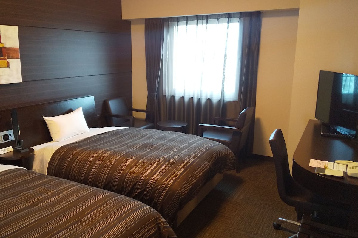 A hotel room with two beds, a desk with a television, a chair, a small table, and a window with curtains.