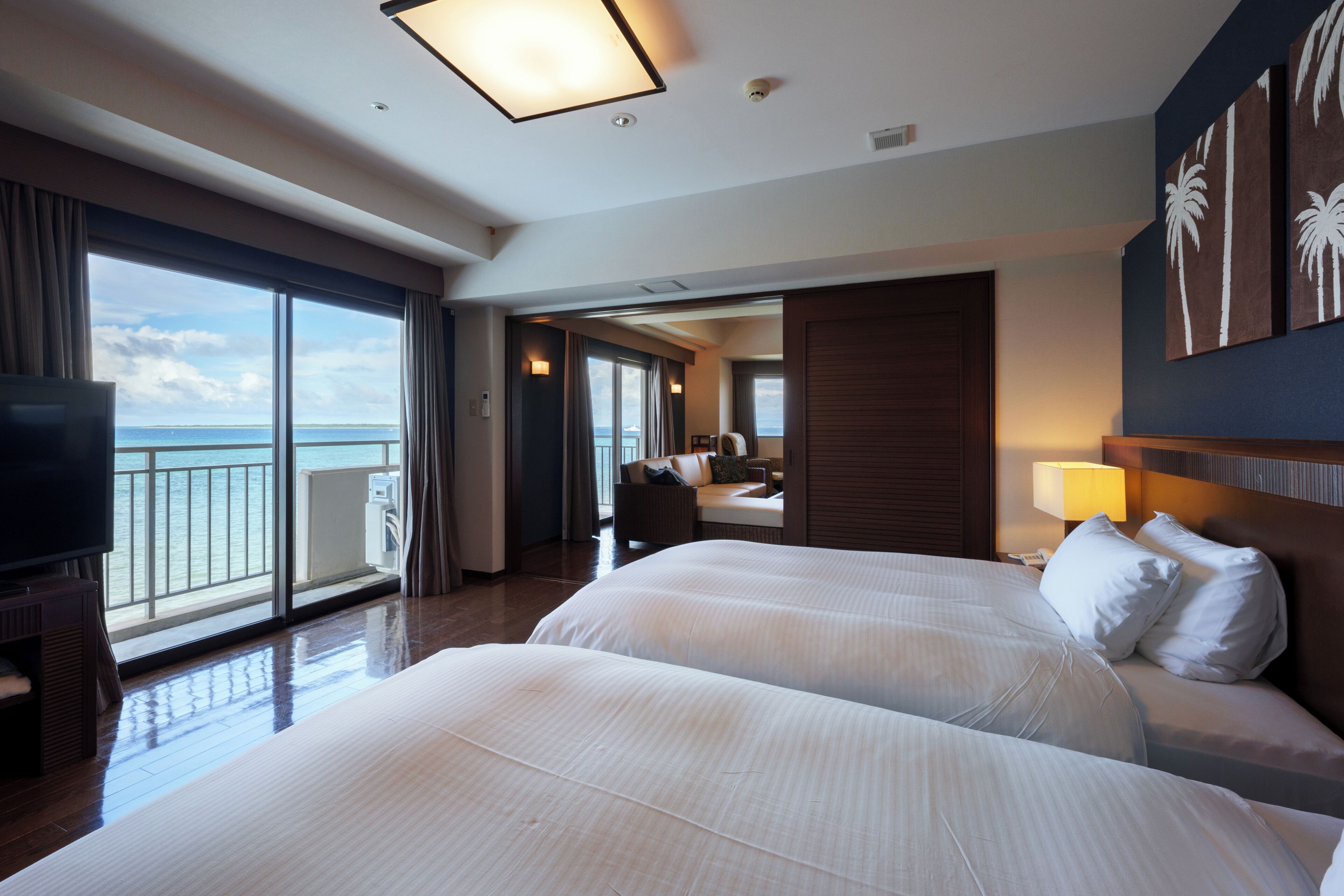 A hotel room with two beds, a TV, a balcony with a view of the sea, and a large window.