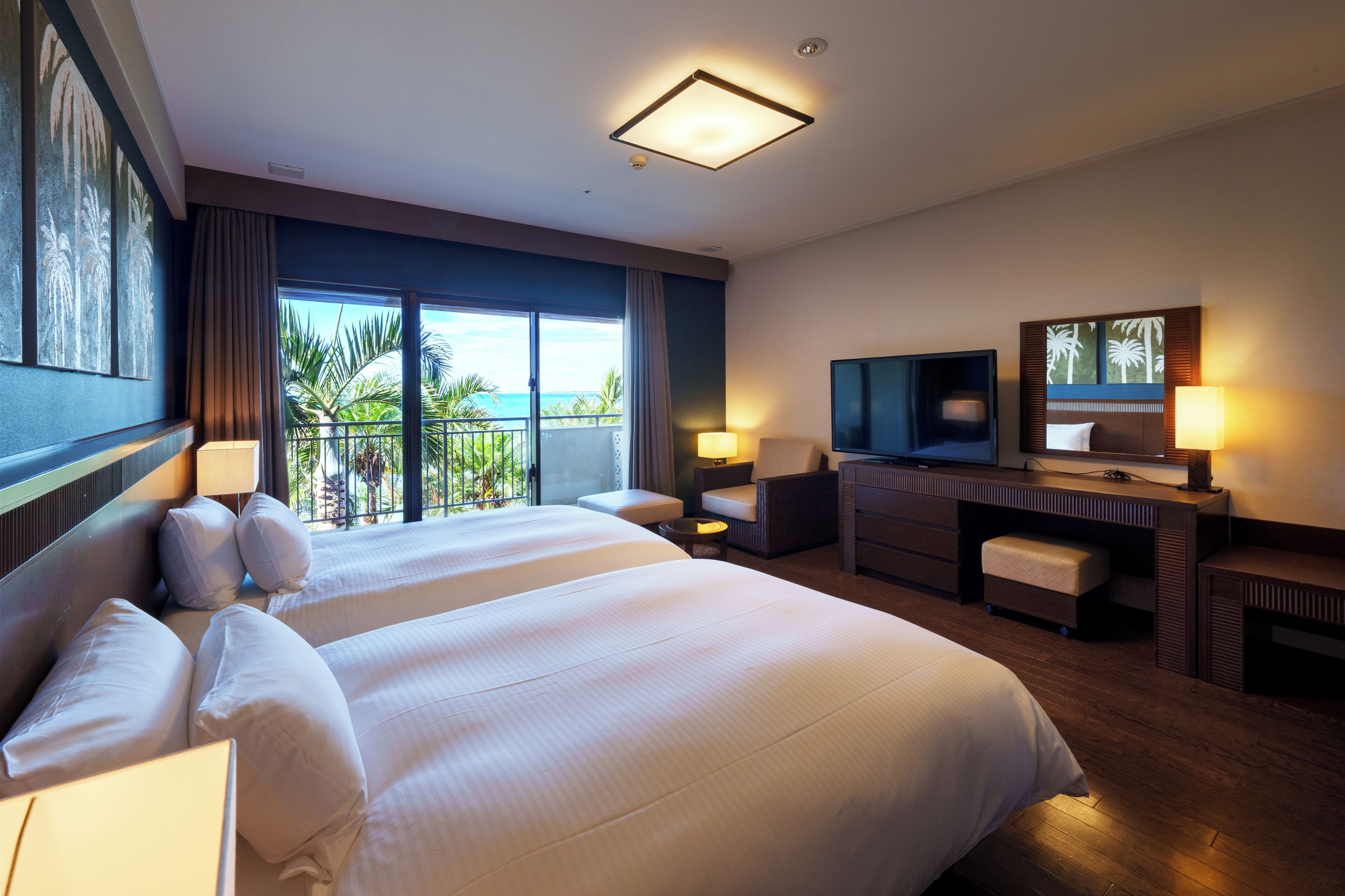 A hotel room with a large bed, a desk with a TV, and a balcony with a view of palm trees.