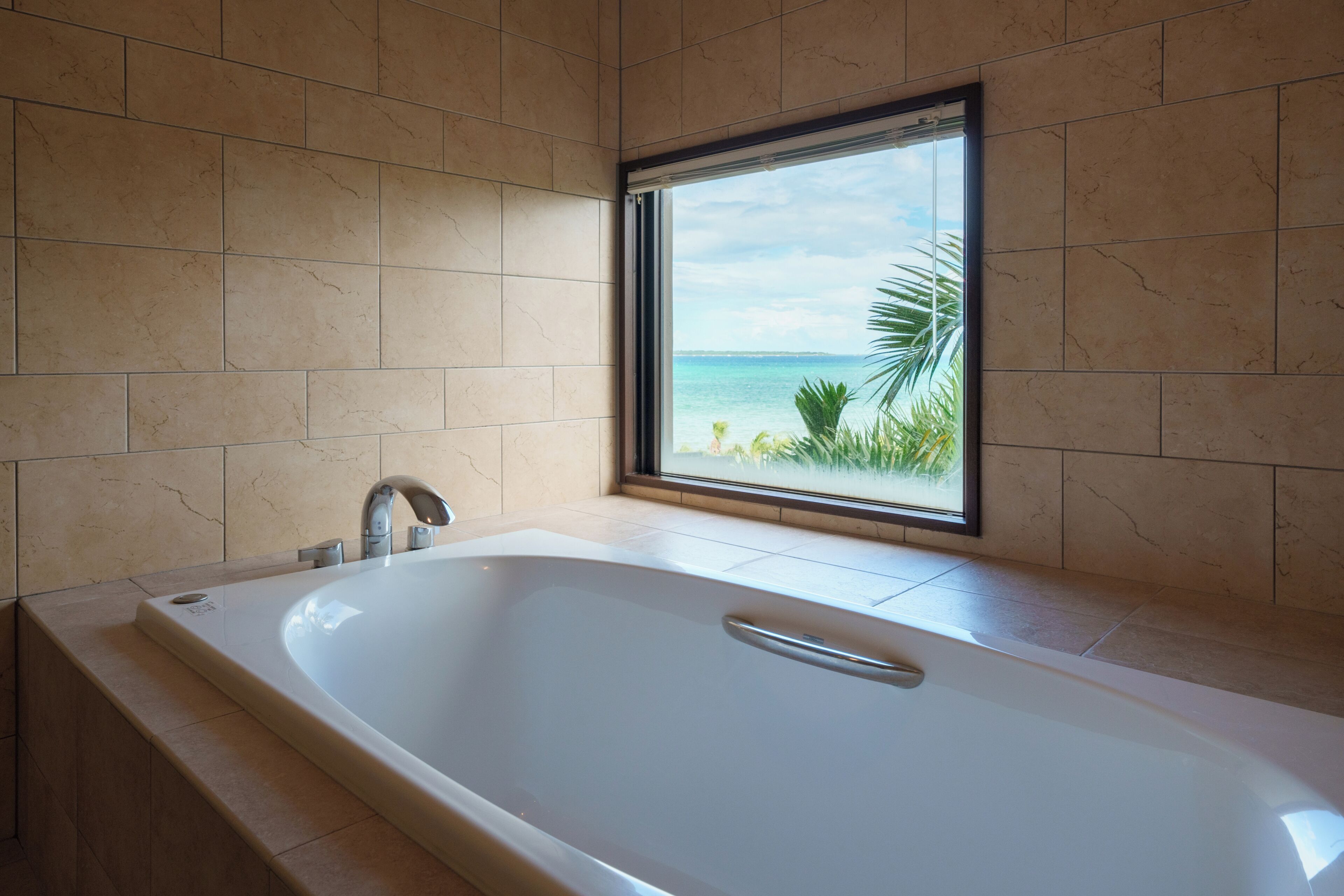 A modern bathroom with a large bathtub, a window with a view of the sea, and tiled walls.