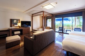 A hotel room with a large bed, a desk with a TV, a wicker sofa, and a balcony with a view of the ocean.