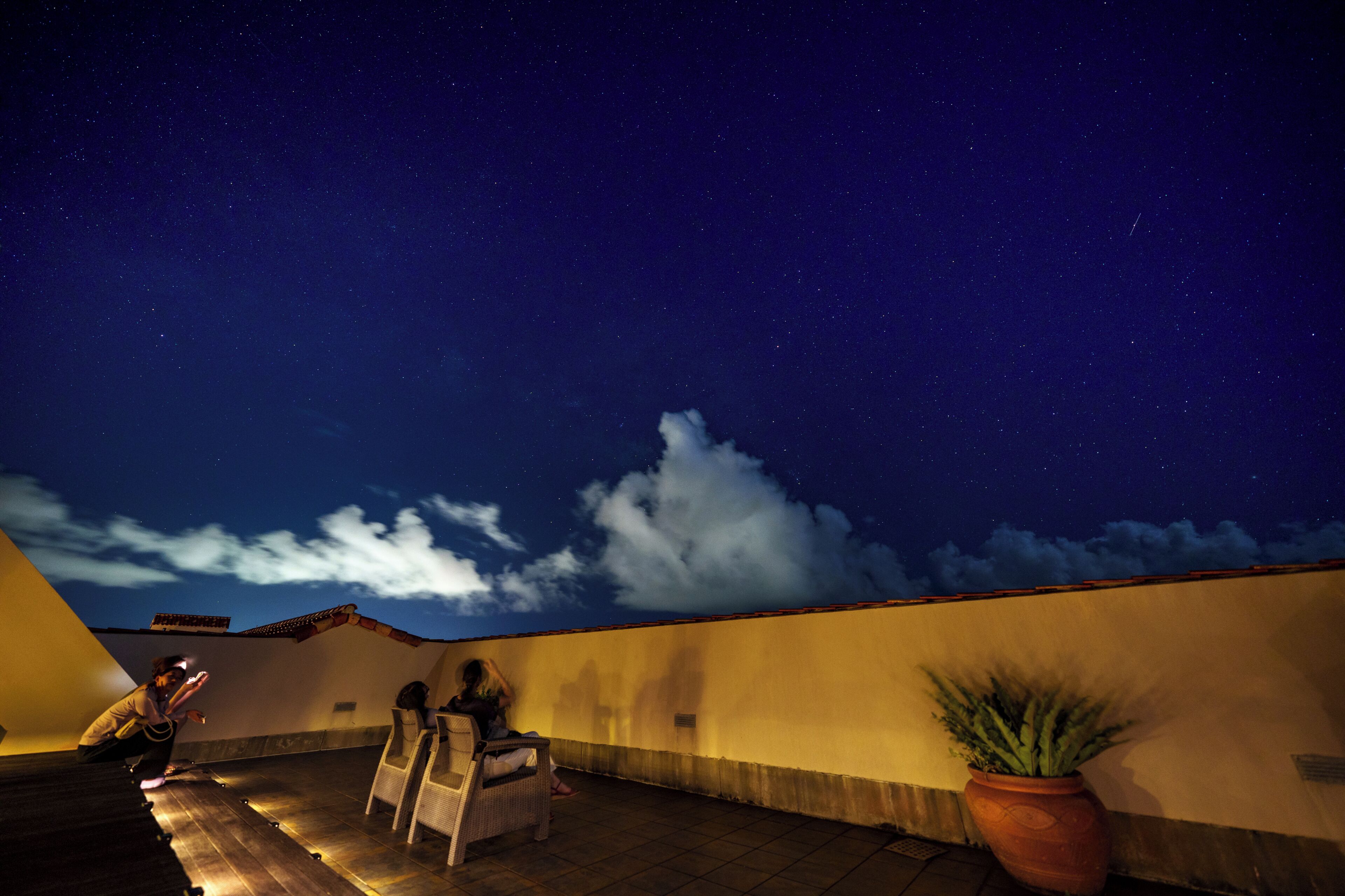 A rooftop terrace with a clear night sky, a potted plant, and a person sitting on a chair.