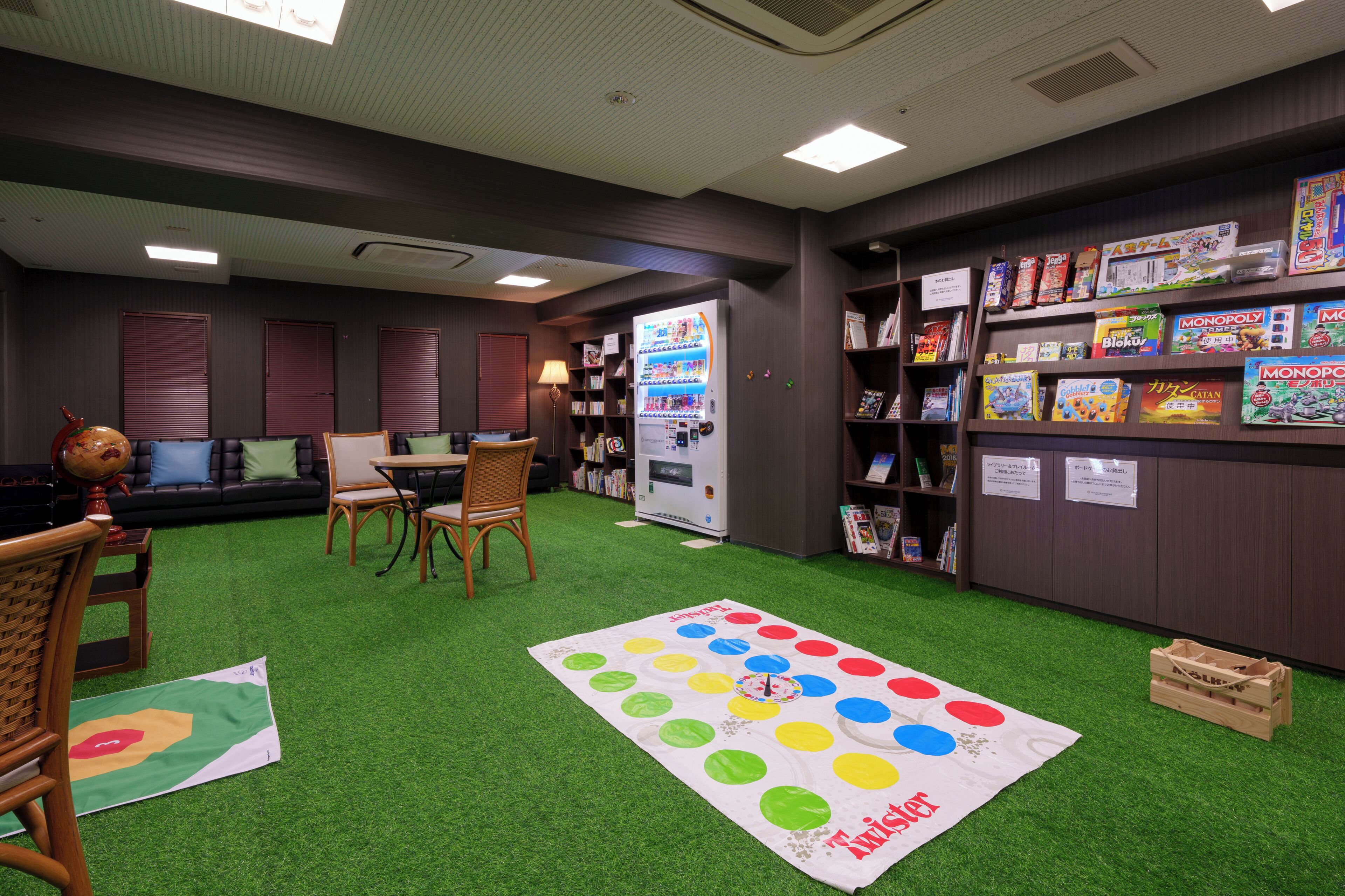 Una habitación con césped artificial verde, un juego de mesa Monopoly y una máquina expendedora.