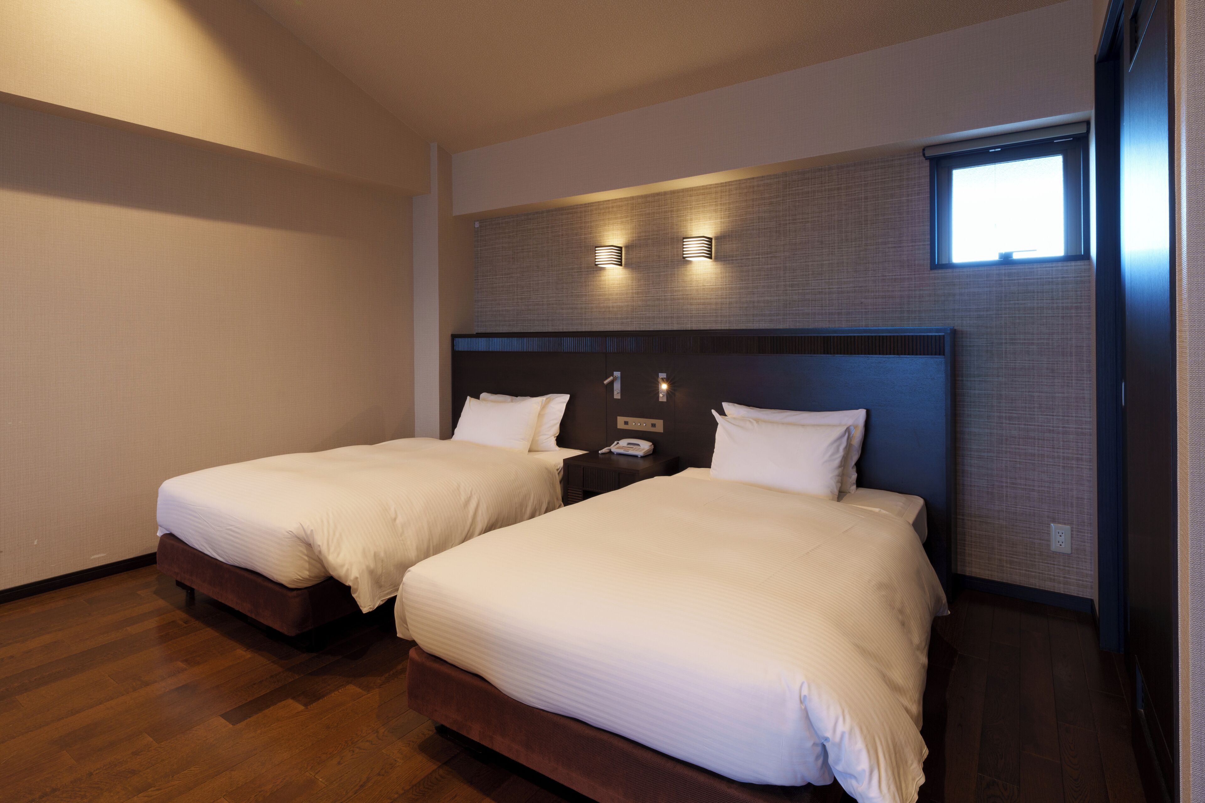 A hotel room with two beds, a wooden floor, a headboard with a light fixture, and a small window.