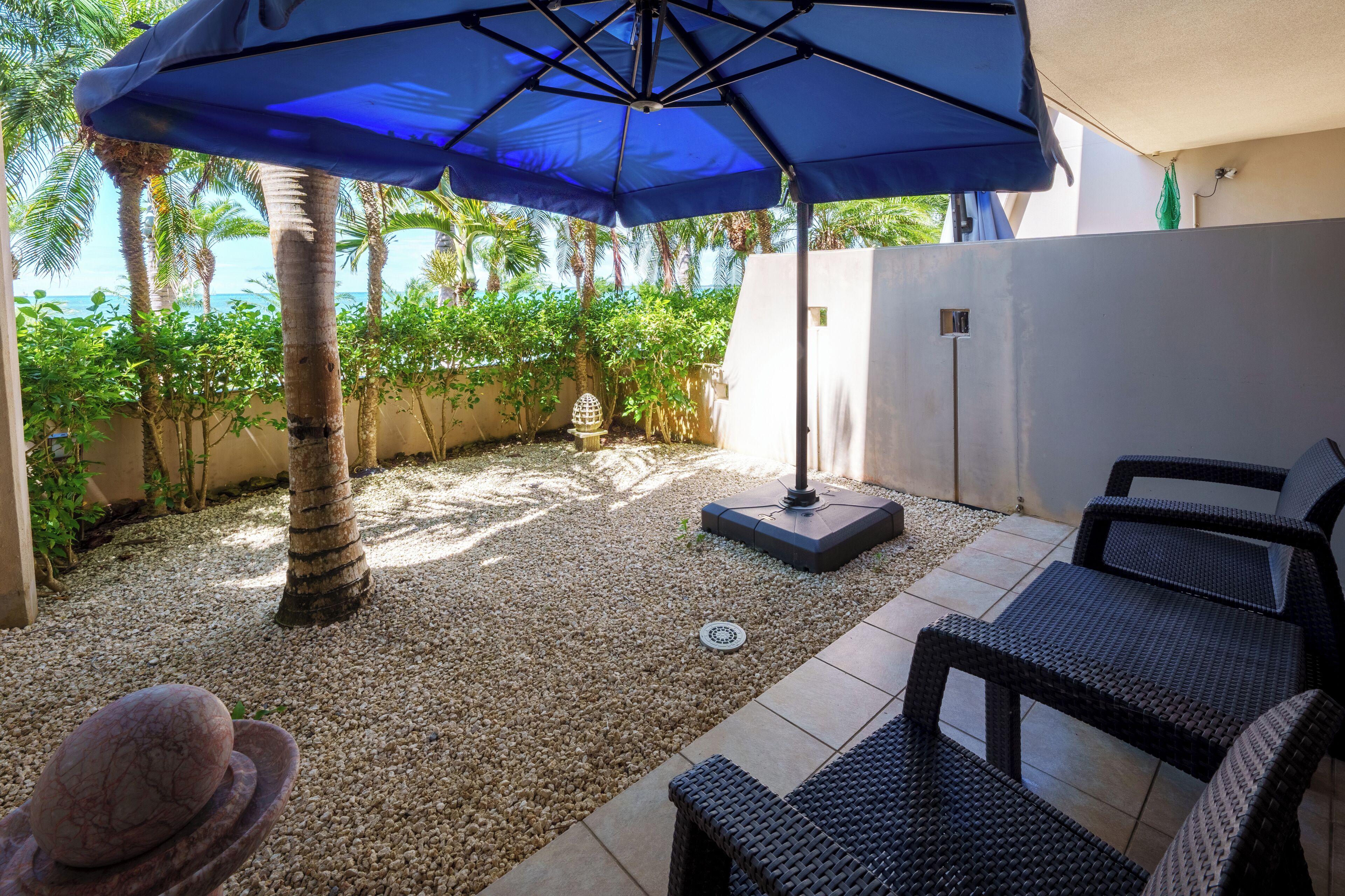 Une terrasse extérieure avec un parasol bleu, des meubles en osier, une sculpture en pierre et une vue sur l’océan.