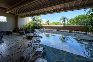 Une piscine en pierre, à l’eau claire, entourée de rochers et surmontée d’une pergola en bois.