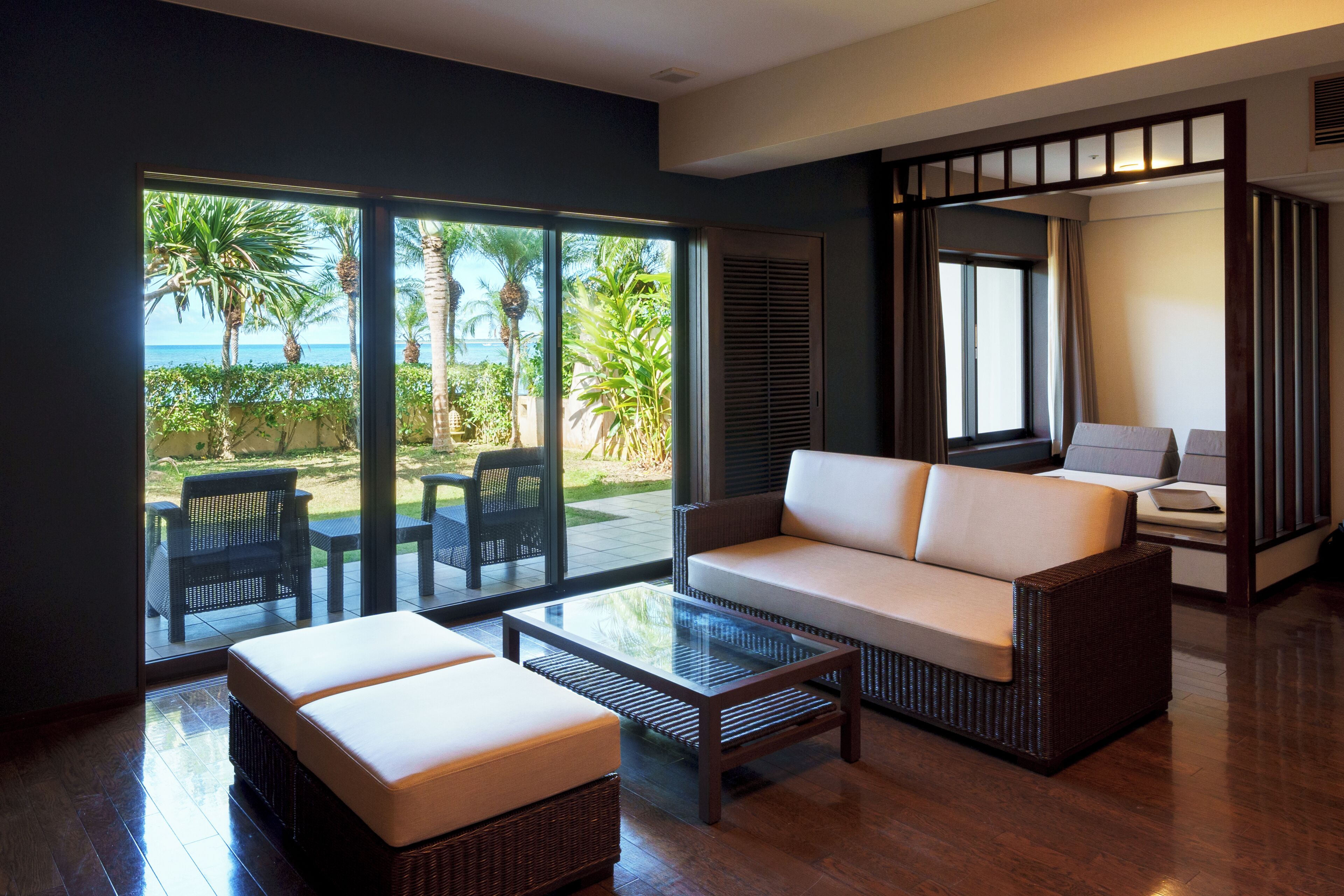 A modern living room with a sofa, ottoman, and coffee table, large sliding glass doors leading to a patio with outdoor furniture and palm trees.